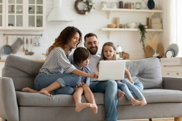 Happy young family with little kids sit on sofa in kitchen have fun using modern laptop together, smiling parents rest on couch enjoy weekend with small children laugh watch video on computer at home