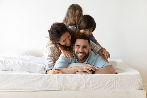 Portrait of happy young parents and small preschooler kids lying on white mattress in bedroom look at camera together, smiling family with little children relax on bed have fun, furniture concept