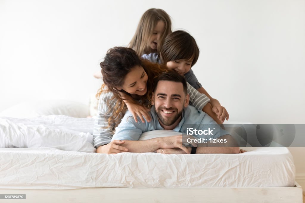 Happy young family with kids relax in bedroom Portrait of happy young parents and small preschooler kids lying on white mattress in bedroom look at camera together, smiling family with little children relax on bed have fun, furniture concept Family Stock Photo