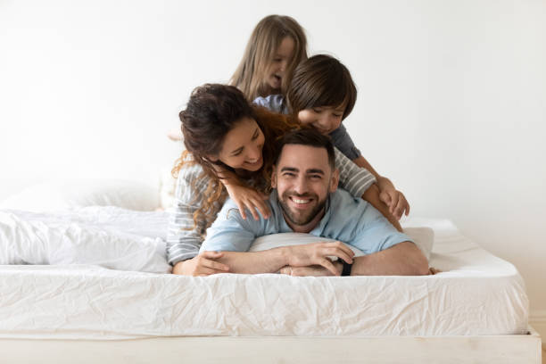 Happy young family with kids relax in bedroom Portrait of happy young parents and small preschooler kids lying on white mattress in bedroom look at camera together, smiling family with little children relax on bed have fun, furniture concept social services photos stock pictures, royalty-free photos & images