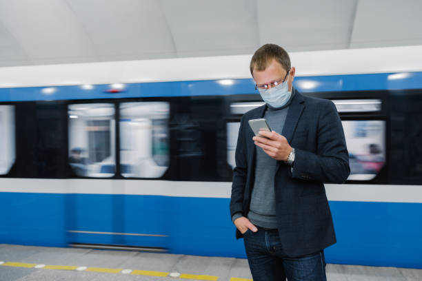 il passeggero maschio indossa pose maschera facciale sulla piattaforma, aspetta il treno, commuta in metropolitana, concentrato nel dispositivo smartphone, si legge sulle notizie online. l'aeità virale era in luogo pubblico. focolaio di coronavirus - feed on foto e immagini stock