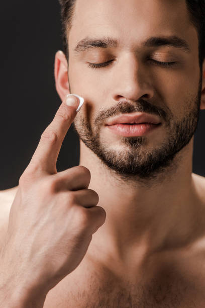 istock naked man with closed eyes applying face cream isolated on black 1215804836