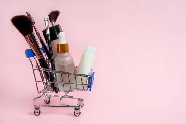 Skincare cosmetics and makeup brushes in a grocery shopping cart on a pink background. Skin care concept