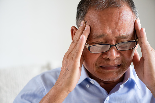 Asian old man sitting on sofa and having a headache at home. Senior healthcare concept.