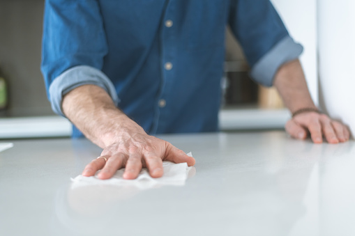 Close-up shot of man using a disinfecting wipe to clean a kitchen counter. The modern kitchen has a white counter. The man is wearing a casual shirt with sleeves rolled up.