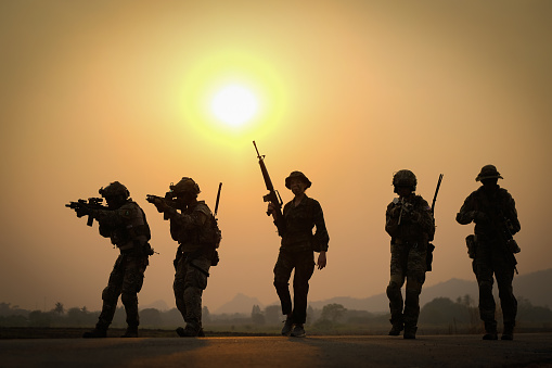 Silhouette of group of soldier hold the weapon and walk along the road with sunset in the back.
