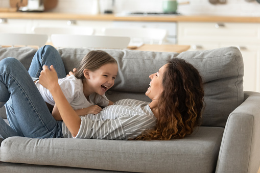 Overjoyed young mother lie on couch in kitchen have fun play with smiling little daughter enjoy weekend at home, happy mom feel playful engaged in childish activity with small preschooler girl child