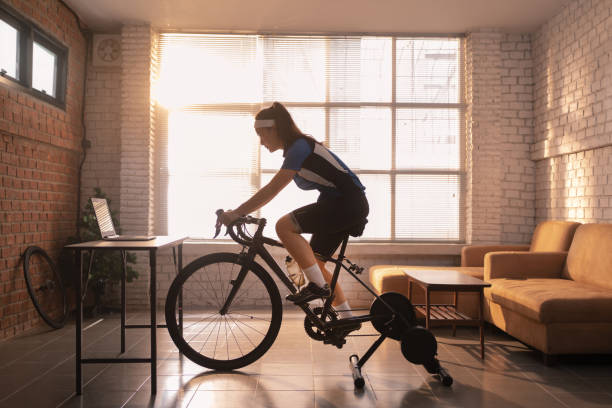 アジアの女性サイクリスト。彼女は家の中で運動している。トレーナーでサイクリングし、オンラインバイクゲームをプレイすることによって - cycling shorts ストックフォトと画像