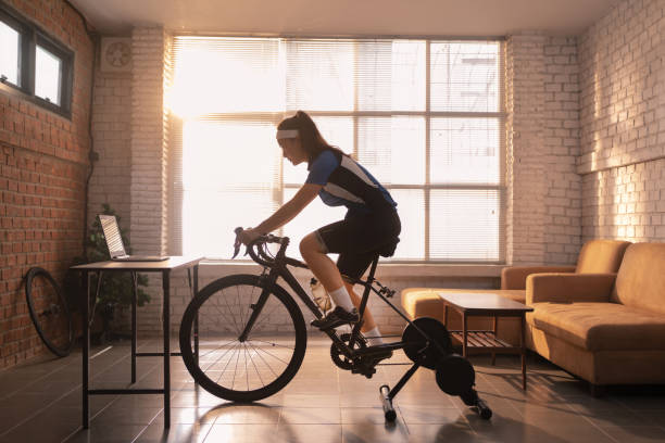 mulher asiática ciclista. ela está se exercitando em casa. pedalando no treinador e jogando jogos online de bicicleta - short cycle - fotografias e filmes do acervo