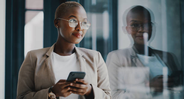 Thinking of the right response Shot of a young businesswoman using a cellphone while standing at a window in an office piece of mind stock pictures, royalty-free photos & images