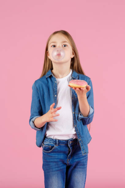 маленькая девочка в солнцезащитных очках взрывает розовую жевательную резинку на светло-фиолетовом фоне - chewing gum bubble blowing little girls стоковые фото и изображения