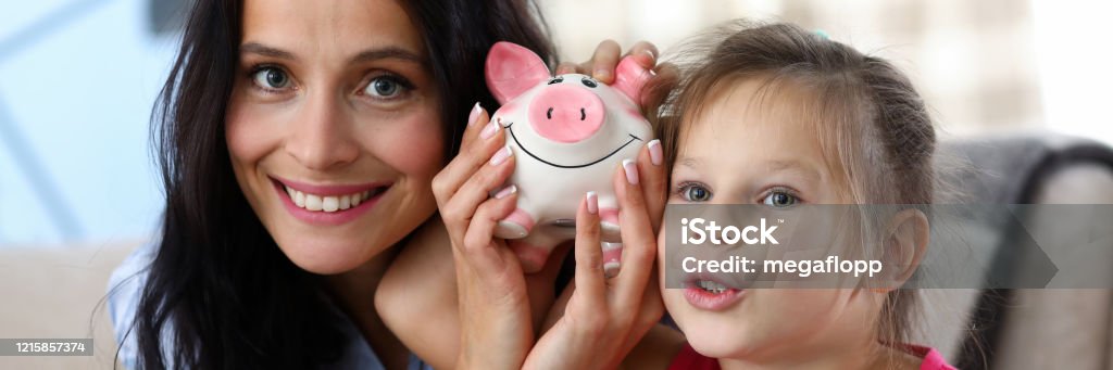 Cheerful parent with child Portrait of happy mother and daughter shaking piggy-bank with coins. Little girl and her mom posing at home. Investment in future and saving money concept Family Stock Photo