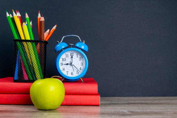 istock The blue alarm clock along with multi-colored pencils stand on a stack of red books, next to an apple 1215864608