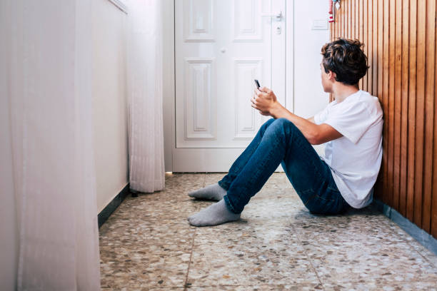 滞在ホームロックダウン緊急コロナウイルスのための家の中の人々 - 若い男はに座ります。ドアの希望を見て電話で床に出て、人生を楽しむために待ちます - on the inside looking out ストックフォトと画像