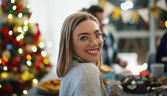Group of friends sitting at the dinning table, eating and drinking, having fun during Christmas times. Young woman at the table is smiling and looking at camera over the shoulder.