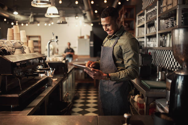 Cheerful young afro-american cafe owner wearing black striped apron using digital tablet Young coffee shop owner holding using digital tablet working while standing behind counter barista photos stock pictures, royalty-free photos & images