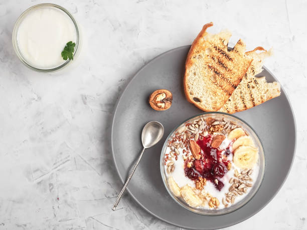 Healthy breakfast concept. Yogurt with granola, cherry jam and nuts in a cup on a gray background. Toast bread and a can with yogurt on the table. Top view, horizontal orientation.