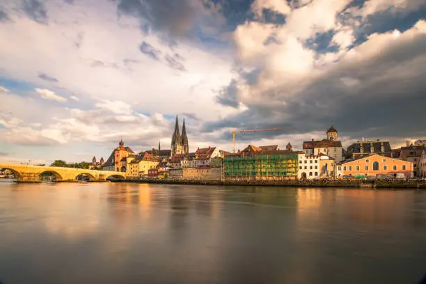 Panoramic high detail view from the Danube river towards  historical Stone Bridge, Bridge tower and iconic architecture buildings in the late afternoon hours, Regensburg, Germany. Long exposure cloudscape photograph with beautiful water reflections. Shot on Canon EOS full frame system.