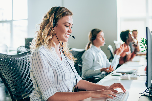Smiling beautiful young businesswoman working in call center. Shot of a adorable young woman working in a call center with her team. Confident female operator is working with colleagues. Call center operators sitting in a row at desks.