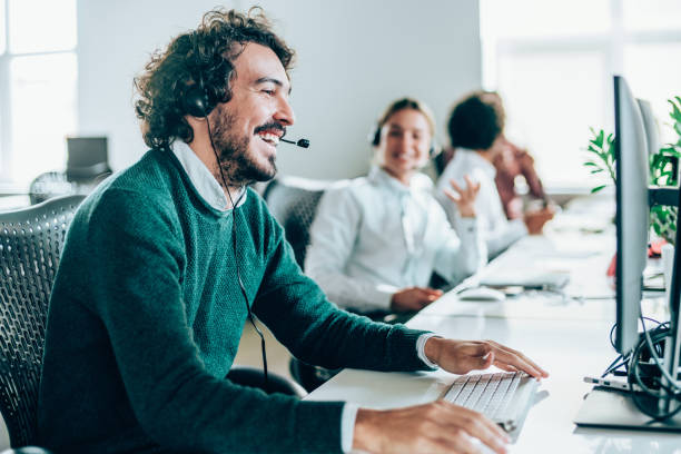 Call center workers. Smiling handsome young businessman working in call center. Shot of a cheerful young man working in a call center with his team. Confident male operator is working with colleagues. Call center operators sitting in a row at desks. service department stock pictures, royalty-free photos & images