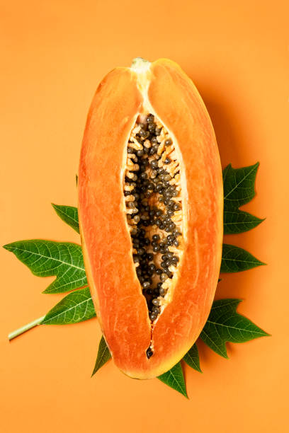 Ripe freshly cut papaya fruit with green papaya leaf on orange background, flat lay, vertical composition stock photo
