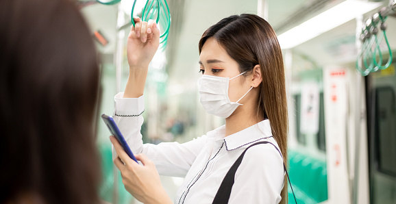 Asian business woman use a smartphone with surgical mask face protection while commuting in the metro or train