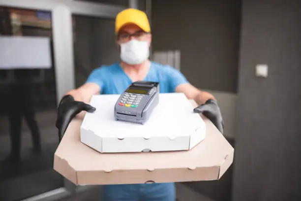 Photo of Deliveryman with protective medical mask holding pizza box and POS wireless terminal for card paying - days of viruses and pandemic, food delivery to your home and safety hygiene measures.