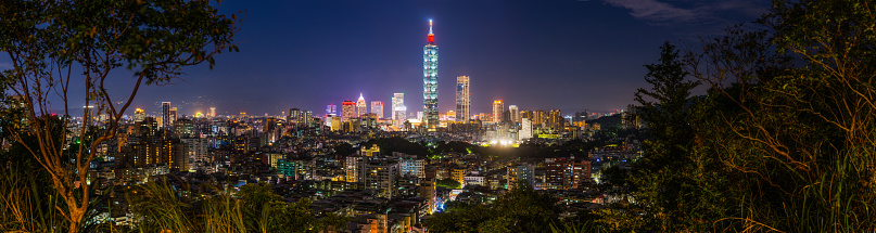 The iconic tower of Taipei 101 and the neon night skyscrapers and crowded cityscape of downtown Taipei framed by the leafy foliage of a nearby mountain park in the heart of Taiwan’s vibrant capital city.