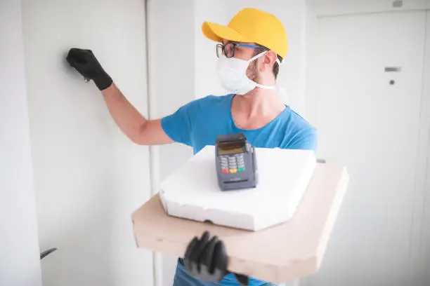 Deliveryman with protective medical mask holding pizza box and POS wireless terminal for card paying, knocking at the door - days of viruses and pandemic, food delivery to your home and safety hygiene measures.