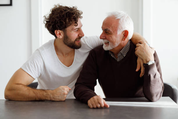 Father and son 70-year-old father is sitting at the table with his 30-year-old son and smiling. social history photos stock pictures, royalty-free photos & images