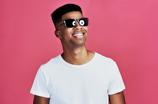 Cropped shot of a handsome young man standing alone and wearing sunglasses against a pink background in the studio