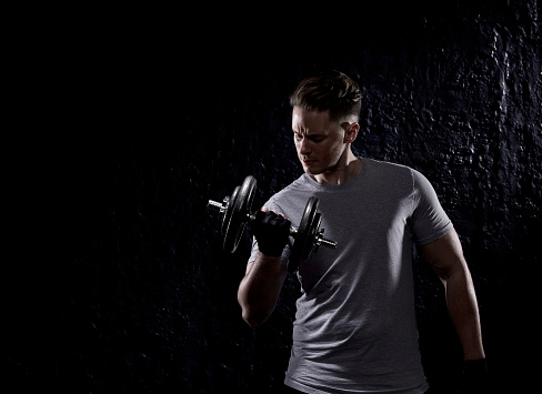 Portrait of an athletic young man with neatly-trimmed hair, photographed against a dark background lifting weights