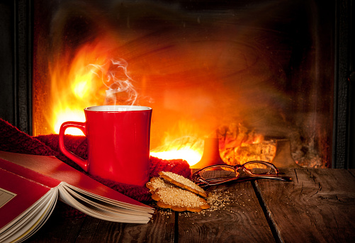 Hot tea or coffee in a red mug, ginger cookies, book and glasses on vintage wood table. Fireplace as background. Christmas or winter warming drink. Layout with free text space.