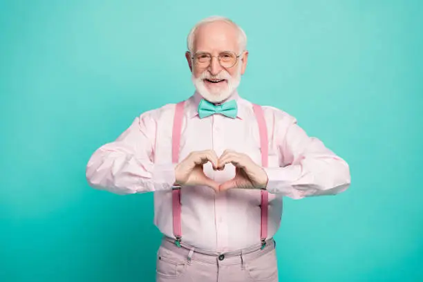 Photo of Photo of attractive grandpa making heart figure fingers hands inviting wife romance date wear specs pink shirt suspenders bow tie trousers isolated teal color background