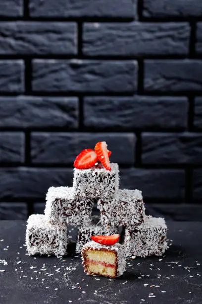 Photo of Australian party food dessert: lamingtons with strawberry jam filling, coated with chocolate and shredded coconut served on a round wire rack on a dark concrete table