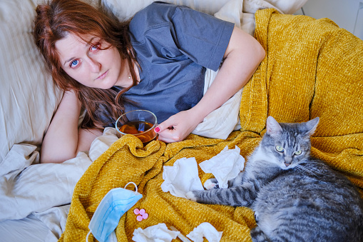 Woman with a coronavirus drinks tea while lying on a bed next to a cat