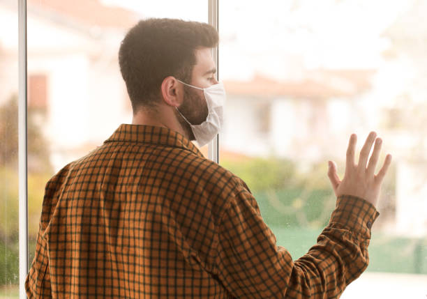 istock Man wearing a protective medical mask looking out of the window during quarantine 1215935535