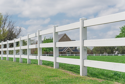 Selective focus on the white fence with blurry two story farm house in background. Long wooden fence along green grass garden under cloud blue sky, farmland ranch in Ennis, Texas, America.