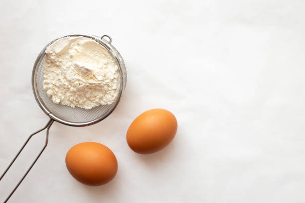 Two eggs and sifter with flour on white parchment paper, top view. Cooking concept with copy space stock photo