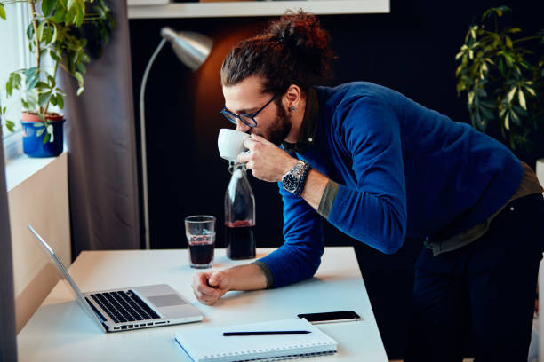 istock Young attractive bearded hipster with curly hair leaning on table, drinking coffee and surfing on internet while standing in home office. Work from home concept. 1215945529