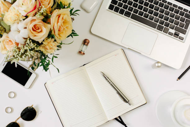 istock Office desk workspace with computer, opened notebook and flowers bouquet and stationery on white table. Flat lay, top view 1215952362