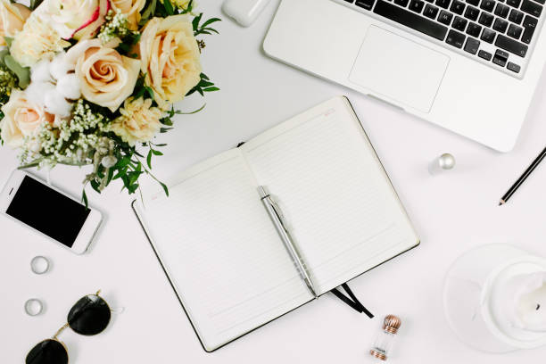 istock Office desk workspace with computer, opened notebook and flowers bouquet and stationery on white table. Flat lay, top view 1215952531