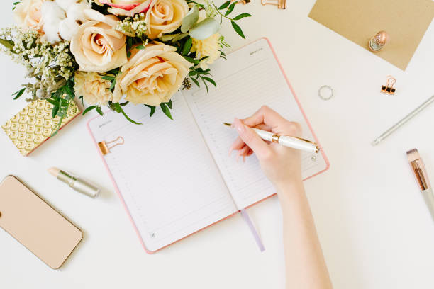 istock Hand wtite in pink opened notebook on white background. Tender mood and blogging concept with beautiful roses bouquet and accesoires in flat lay style 1215952687