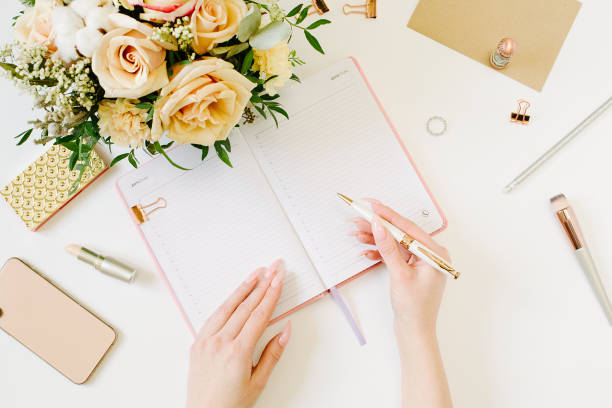 istock Female hands wtite in pink opened notebook on white background. Tender mood and blogging concept with beautiful roses bouquet and accesoires in flat lay style 1215953788