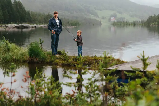 July 7, 2012 - Norway: Little cute boy, toddler, is holding a fishing rod and trying to catch a fish while his father stands beside him smiling. Lake in Norway, family togetherness.