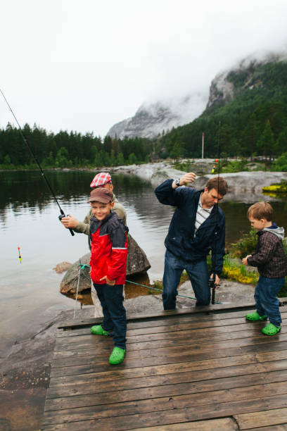 istock Cute handsome boy is smiling, looking at camera hands in pockets while his father, grandfather and brother are preparing fishing rods on an overcast day in Norway at a lake and mountains 1215955771