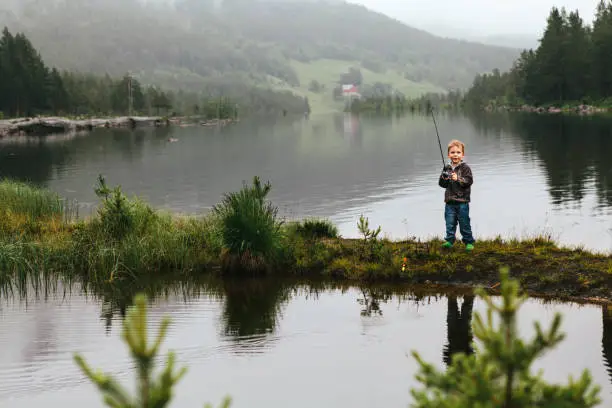 July 7, 2012 - Norway: beautiful little boy is standing excited on a small strip of grass on a mountain lake, holding a fishing rod and looking at camera smiling.