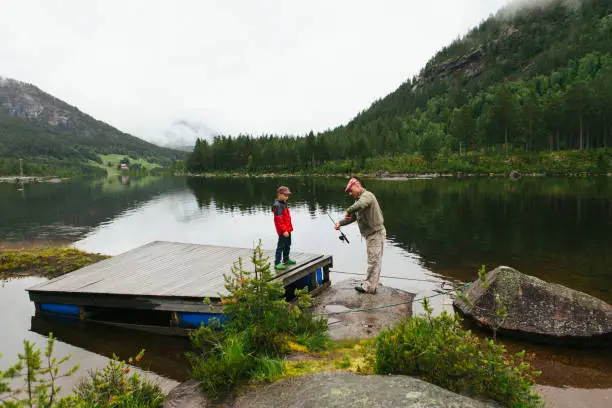 July 7, 2012 - Norway: grandson is learning fishing from his grandfather - looking down at the fishing rod while grandpa is explaining. Beautiful lake near the mountain in Norway