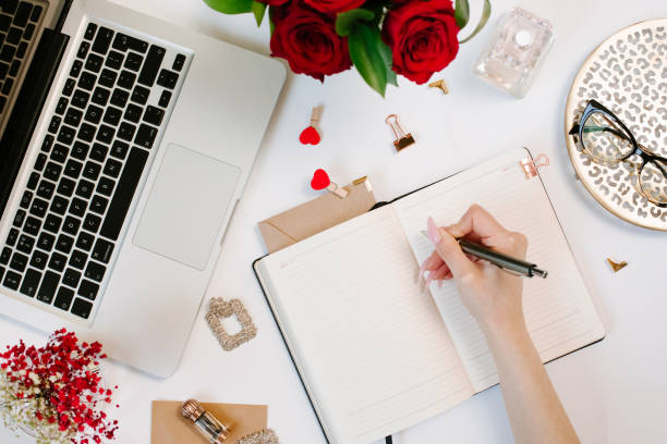 istock Female hand write in notebook. Office desk workspace with computer, roses, glasses. Flat lay style. Freelance and remote work concept 1215958538