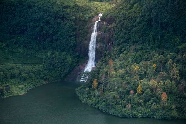 A waterfall in a mountainous landscape. The waterfall is located in the center of the image, with water cascading down the rocks. The landscape is covered in greenery, with trees and bushes covering the hills. The sky is blue with some clouds, and the mountains in the background are covered in mist. There are some buildings visible on the right side of the image, possibly a village or town. The photograph is taken from a high vantage point, looking down on the landscape.